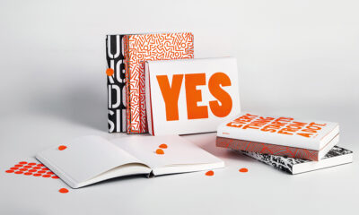Eine Sammlung von Notizbüchern mit grafischen Mustern in Schwarz, Weiß und Orange, darunter eines offen mit der Aufschrift 'YES' auf der Vorderseite, umgeben von kleinen runden orangenen Aufkleberchen