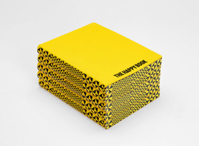 Stack of yellow notebooks titled 'THE HAPPY BOOK' with a graphic black-and-yellow pattern on the sides