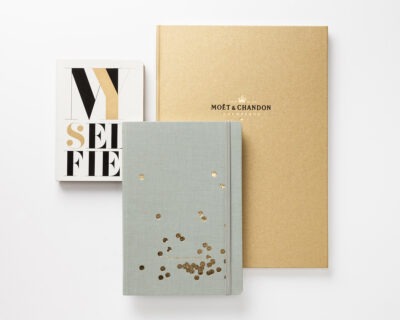 Collection of notebooks, including one with 'My Selfie' inscription, a golden one with Moët & Chandon branding, and a light gray linen book with golden accents