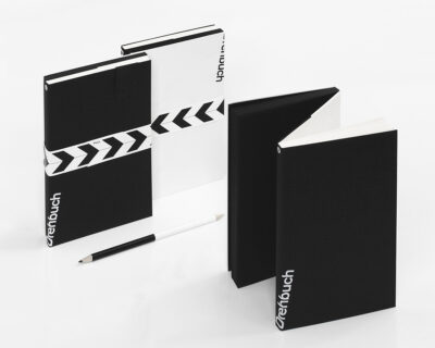 Carnets noir et blanc avec le branding 'Drehbuch', l'un avec une couverture noire et l'autre avec un design à rayures diagonales