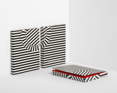 Two black and white striped notebooks with a circular cutout design, standing, and another striped notebook with red accents, lying down