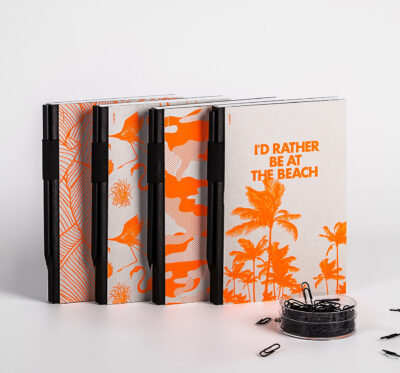 Notizbücher mit tropischem Design in Orange und Weiß mit dem Aufdruck 'I'D RATHER BE AT THE BEACH', neben Behälter mit Büroklammern