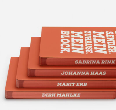 Stapel orange-roter Notizbücher mit Namenspersonalisierung auf den Buchblockkanten und dem Titel 'MEIN ZUHAUSE MEIN BLOCK' in Weiß auf dem Cover