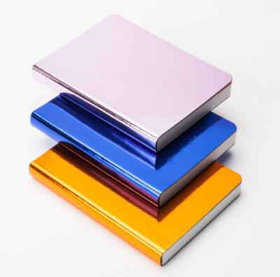 Stapel von metallisch schimmernden Notizbüchern in Rosa, Blau und Orange