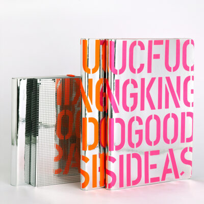 Notizbücher in verschiedenen Größen und Designs aus spiegelndem Einbandmaterial, eines mit orangener Schrift und eines mit pinker mit der Aufschrift 'FUCKING GOOD IDEAS' auf dem Cover