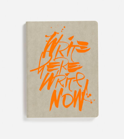 Notizbuch mit natürlicher, grauer Papierstruktur und leuchtend orange-graffitiartiger Aufschrift 'WRITE HERE WRITE NOW' auf der Vorderseite
