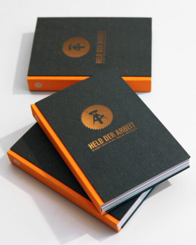Schwarze Notizbücher mit orangenem Seitenrand und 'Held der Arbeit' Prägung auf dem Cover